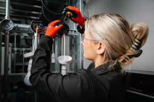 Wearing red gloves and protective wrap around glasses, a student with tied up blonde hair faces away from us and works on cabling of a few hanging lightbulbs attached to metal framing